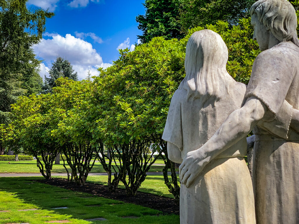 A statue viewed from behind of a man and woman with their arms around each other with grass, trees and rhododendrons in the background. 