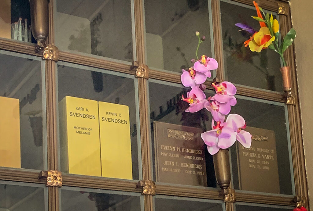 Niches behind glass, some with boxes and names on them and some empty. Orchids placed in flower holder in front. 