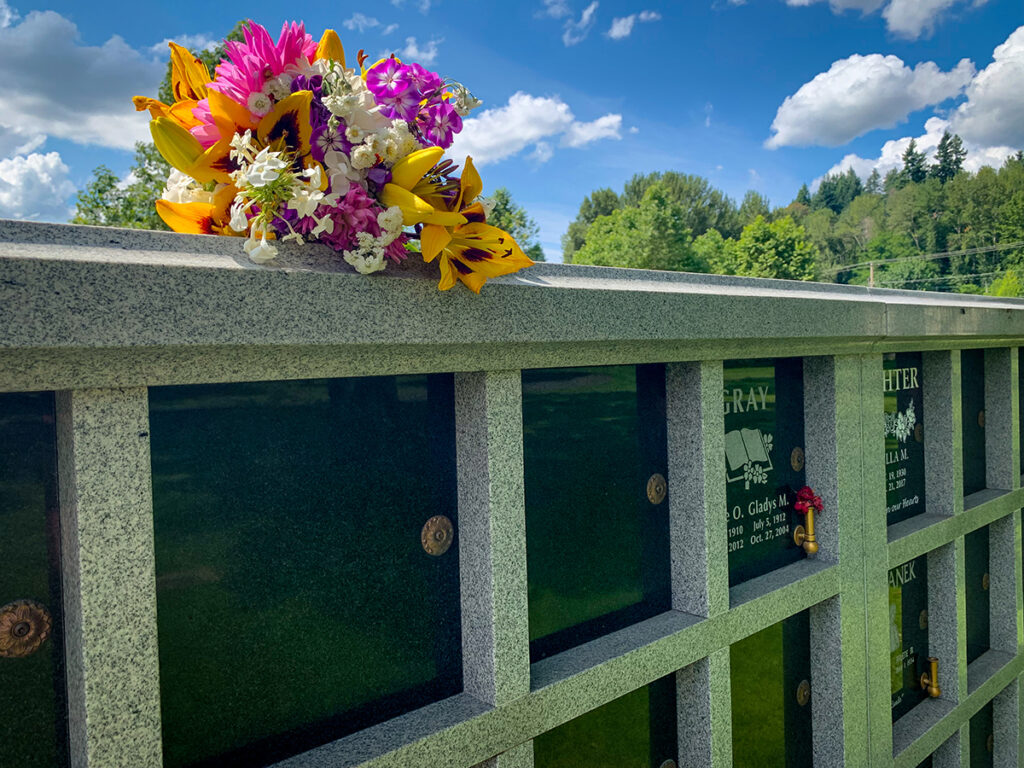 Black granite niches, some etched in white and some blank with a bouquet of pink and yellow flowers on top of the wall and blue sky behind. 